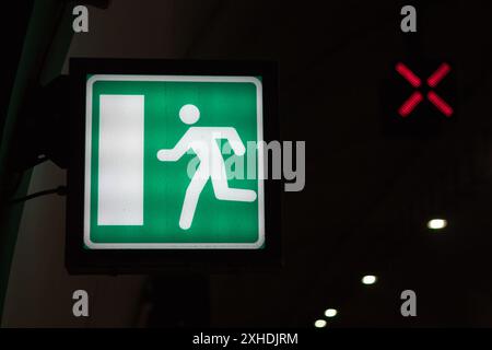 A green exit sign with a white running figure is illuminated in a dark space, with a red X visible above it. Stock Photo