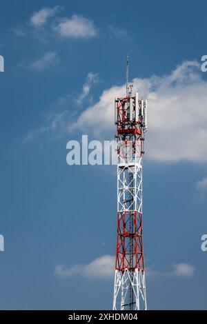 A tall red and white cell tower rises against a bright blue sky dotted with fluffy white clouds. Stock Photo