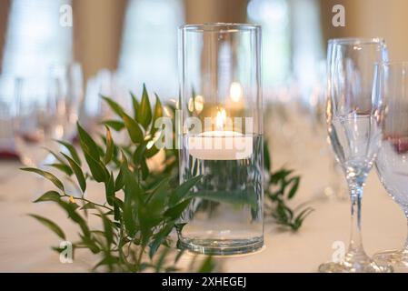 Tall glass vase with lit tea light candle floating inside on wedding reception dinner table with green plants Stock Photo