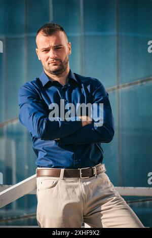 A man with arms crossed, wearing a blue button-down shirt and khaki pants, stands in front of a modern glass building. Stock Photo