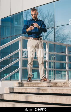 A man in a blue shirt and sunglasses stands on steps outside a modern building, using his smartphone. Stock Photo