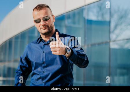 A man wearing sunglasses and a blue button-down shirt gives a thumbs-up in front of a modern glass building. Stock Photo