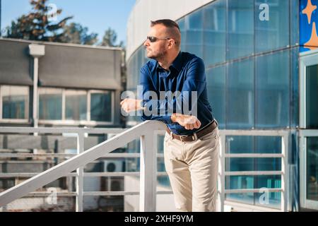 A man wearing sunglasses and a blue shirt leans on a railing outside of a modern building with glass windows. Stock Photo