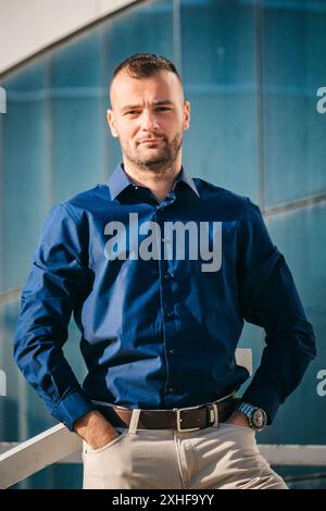 A man in a blue shirt with a serious expression stands in front of a modern glass building. Stock Photo