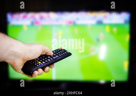 A hand holds a remote control in front of a television screen showing a soccer game. Stock Photo