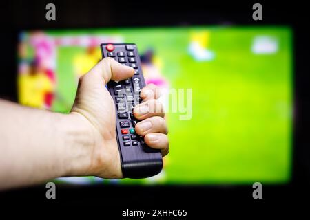 A hand holds a black TV remote control in front of a television screen displaying a blurry sports game. Stock Photo