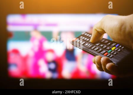 A hand presses a button on a black television remote control, with a blurred television screen in the background. Stock Photo