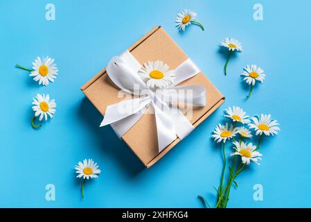 Gift box with white ribbon and Chamomile flowers on blue pastel background, top view. Mothers Day, Fathers Day, Birthday design concept. Stock Photo