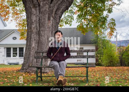 A happy chinese woman sitting on a park bench under a maple tree in manchester vermont in autumn. Stock Photo