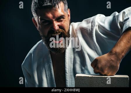 Angry karate man showing punch and breaking concrete brick with fist. Screaming male mixed martial arts fighter smashes concrete slab with his hand. B Stock Photo