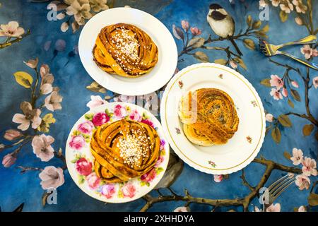 Three plates of classic Swedish yeast pastries sit on a blue patterned tablecloth decorated with blossoms and a bird: kanellbullar (cinnamon buns) topped with pearl sugar, and kardemummaknutar (cardamom knots) sprinkled with poppy seeds and pearl sugar. Both pastries rank among the most popular treats of Swedish fika, the extended coffee break. Two silver forks lie ready beside them. Stortorget, Stockholm, Stockholm County, Sweden. Stock Photo