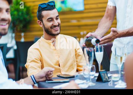 Diverse group of friends dining out at trendy restaurant. Young man in yellow shirt smiling as waiter pours wine. Casual social gathering with multicu Stock Photo