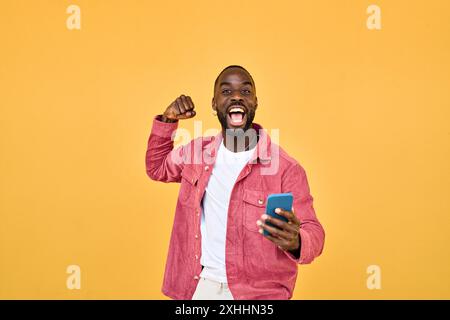 Happy Black man winner holding smartphone celebrating win isolated on yellow. Stock Photo