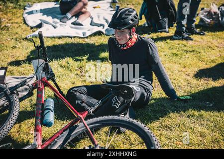 A young man resting on grass with a mountain bike, wearing a helmet and gloves, in a sunny outdoor setting Stock Photo