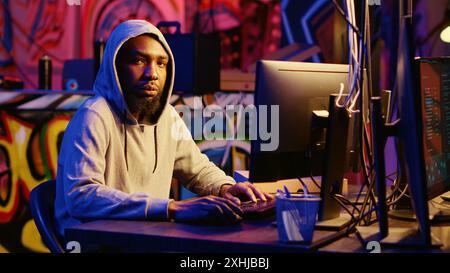 Portrait of african american hacker in abandoned warehouse using phishing technique that tricks users into revealing sensitive information. Hooded cybercriminal stealing passwords Stock Photo