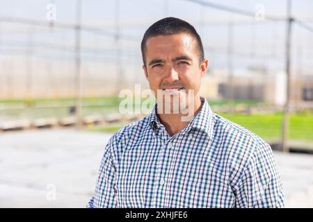 Portrait of positive young male worker posing at greenhouse on background with plantation of sprouts Stock Photo