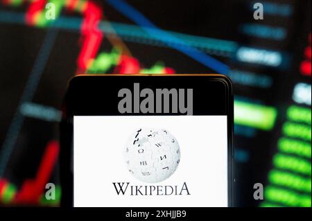 In this photo illustration, the multilingual, web-based, free encyclopedia Wikipedia logo seen displayed on a smartphone with an economic stock exchange index graph in the background. Stock Photo