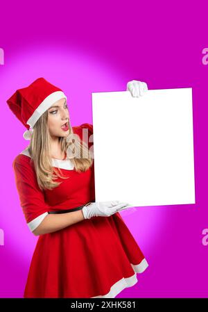 Young blonde woman dressed as Santa Claus looking in amazement at a white sign with copy space isolated on magenta background Stock Photo