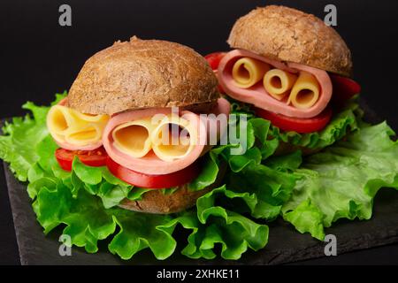Round baguette sandwich with cheese, ham, tomatoes and salad served on black slate serving board on black table Stock Photo