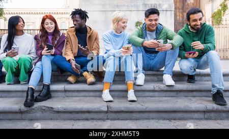 Multiethnic group of young adults sitting together on steps, using mobile phones. Friends enjoying social media and technology outdoors. Diverse mille Stock Photo