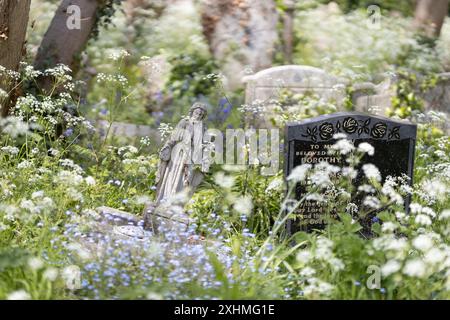 A small, carved stone Jesus, falling over, on a grave amongst a profusion of spring wild flowers and other grave stones. Cemetery, London, UK Stock Photo