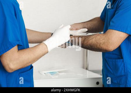 Two healthcare workers in blue scrubs putting on white surgical gloves, preparing for a procedure Stock Photo
