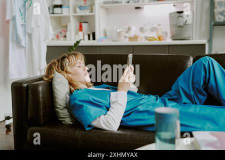 Young Caucasian nurse using smartphone while lying on black leather sofa in on call room Stock Photo