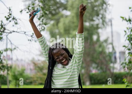 Happy young woman screams WOW holding smartphone jumping on grass in city park. African American with dreadlocks expresses astonishment due to good news. Lady makes energetic moves experiencing joy Stock Photo