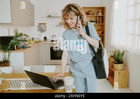 Young happy Caucasian nurse having phone call while reading pateints medical tests via laptop in home kitchen Stock Photo