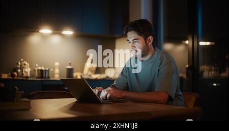 Handsome Authentic Latin Man Sitting at a Table in a Cozy Kitchen and Typing on Laptop Computer at Home at Night. Male Adult Thinking While Browsing the Internet, Checking Videos on Social Networks. Stock Photo