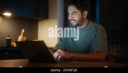 Handsome Authentic Latin Man Sitting at a Table in a Cozy Kitchen and Using Laptop Computer at Home at Night. Male Adult Smiling and Chatting on Social Media, Doing Online Shopping, Checking the News Stock Photo