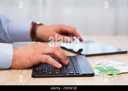 Unrecognizable businessman is typing on a keyboard while is touching tablet screen with his finger. There are several bills on the table. Stock Photo