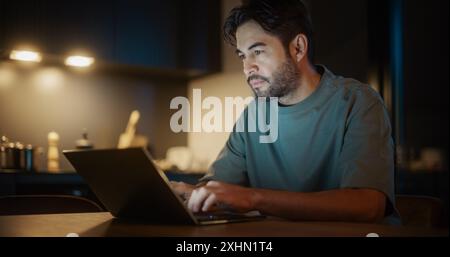 Handsome Authentic Latin Man Sitting at a Table in a Cozy Kitchen and Typing On Laptop Computer at Home at Night. Male Adult Smiling and Chatting on Social Media, Shopping Online, Checking the News. Stock Photo