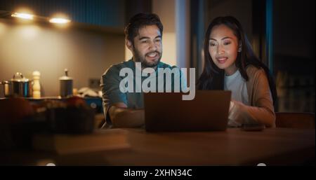 Portrait of Young Couple Using Laptop Computer While Sitting in the Kitchen at Home. Boyfriend and Girlfriend Doing Online Shopping to Renovate the House, Browsing the Internet to Compare Sales Stock Photo