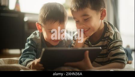 Portrait of Two Male Asian Kids Using a Digital Tablet During Weekend in their Room. Two Cute Brothers Watching Cartoons Together, Focusing on the Screen, Sharing the Gadget and Enjoying their Time Stock Photo