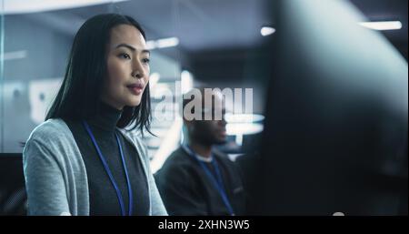 Beautiful Asian Female Working on a Desktop Computer in a Cyber Security Software Development Department. Positive Brunette Female Lightly Smiling While Working on a Project Stock Photo