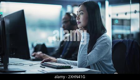 Beautiful Young Asian Female Specialist Working on Computer Together with Professional Colleagues. Data Protection Center with Server Facility, Storage Hardware and Corporate Science Atmosphere Stock Photo