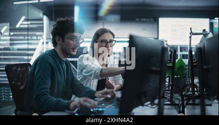 Two Young Colleagues Working on Computers and Talking at a Workplace. Female and Male Software Developers Discussing a Solution for Their Collaborative Artificial Intelligence Project Stock Photo