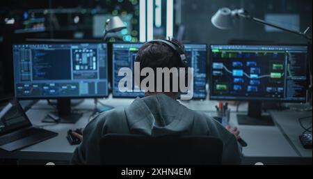 System Administrator Working in a Dark Research Facility on a Computer with Multiple Displays. Software Developer in Casual Clothes Wearing Headphones and Updating Server System Database Stock Photo