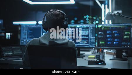 System Administrator Working in a Dark Research Facility on a Computer with Multiple Displays. Software Developer in Casual Clothes Wearing Headphones and Updating Server System Database Stock Photo
