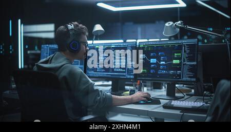 System Administrator Working in a Dark Research Facility on a Computer with Multiple Displays. Software Developer in Casual Clothes Wearing Headphones and Updating Server System Database Stock Photo