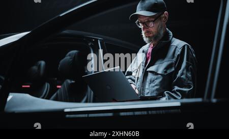 Portrait of a Dealership Car Mechanic Analyzing Diagnostics Results on a Modern Electric Vehicle. Software Developer Working on Laptop Computer, Investigating Settings on a Modern Sustainable EV Stock Photo