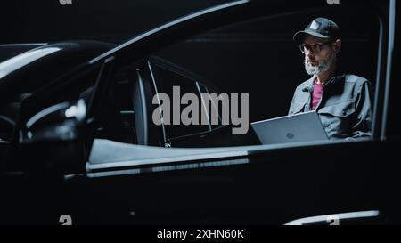 Portrait of a Dealership Car Mechanic Analyzing Diagnostics Results on a Modern Electric Vehicle. Software Developer Working on Laptop Computer, Investigating Settings on a Modern Sustainable EV Stock Photo
