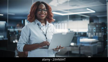 Portrait of a Black Diverse Female Wearing Glasses, Using Laptop Computer, Looking at Camera and Smiling. African Information Technology Specialist, Software Engineer or Developer Stock Photo