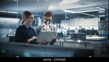 Portrait of Two Creative Young Female and Male Engineers Using Laptop Computer to Analyze and Discuss How to Proceed with the Artificial Intelligence Software. Standing in High Tech Research Office Stock Photo