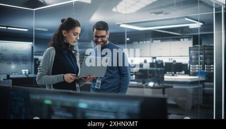 Portrait of Two Happy Female and Male Engineers Using Laptop Computer to Analyze and Discuss How to Proceed with the Artificial Intelligence Software. Casually Chatting in High Tech Research Office Stock Photo