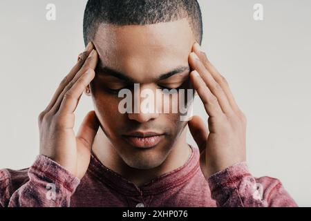 Pained young man, pressing his fingers to his temples while grimacing. Dressed in a maroon shirt, his expression suggests a headache or mental strain, Stock Photo