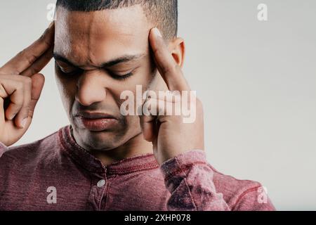Individual with a strained expression, fingers pressed to his temples. Wearing a maroon shirt, he looks to be in pain, possibly from a headache or sev Stock Photo