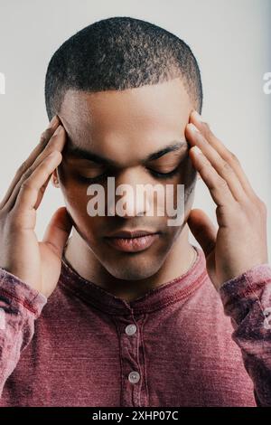 Young man with a pained expression, pressing his fingers to his temples. Wearing a maroon shirt, he appears to be experiencing a headache or intense s Stock Photo