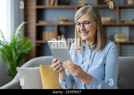 Smiling senior woman using digital tablet at home, sitting on sofa. Enjoying technology, staying connected with family and friends. Modern living room with natural light, bookshelf, and green plant. Stock Photo